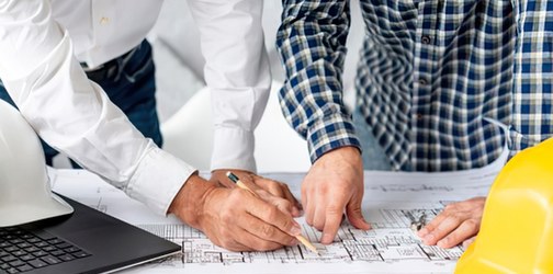 Two project leads — one in a white shirt with a hard hat, one in a blue check shirt — leaning over architectural plans on a worktable, pens in hand, reviewing the details together.